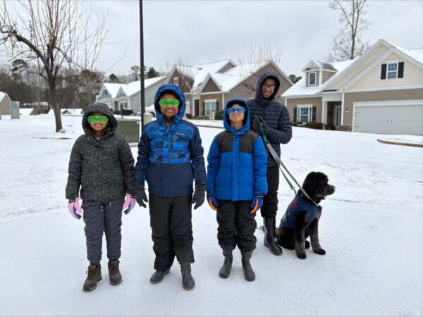 3 children wearing colourful plastic glasses, dad wearing glasses and black pet dog standing out on the yard with snow on the ground