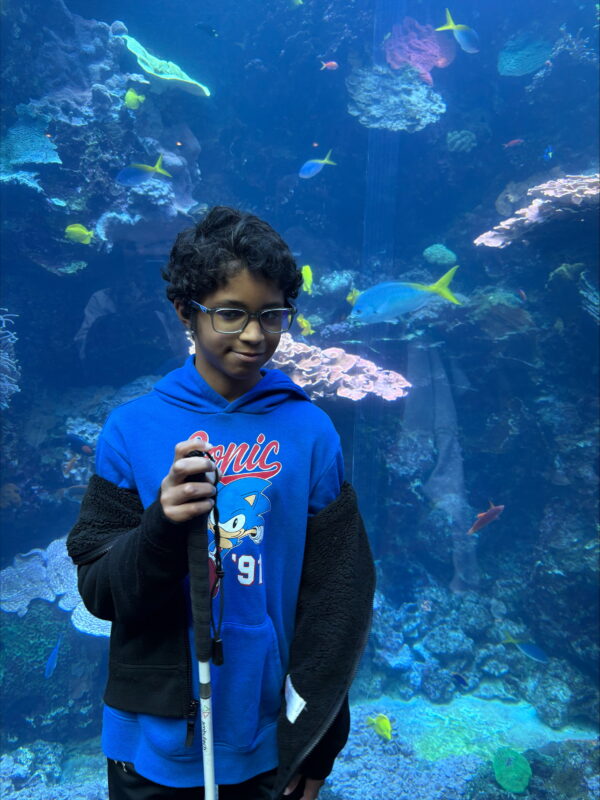 A smiling boy with a white cane in an aquarium, wearing a blue sonic hoodie and blue glasses