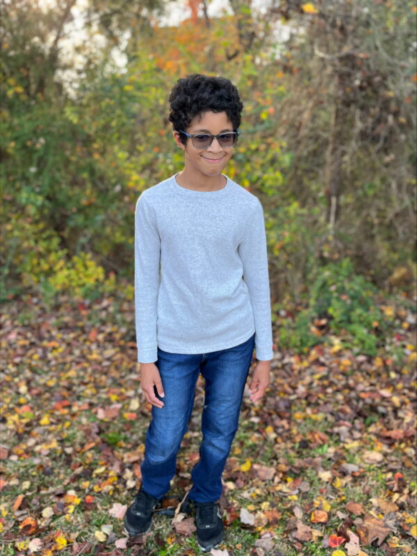 A young boy with blue jeans and gray long sleeve wearing glasses with dark shades standing on the grass with colourful leaves on the ground